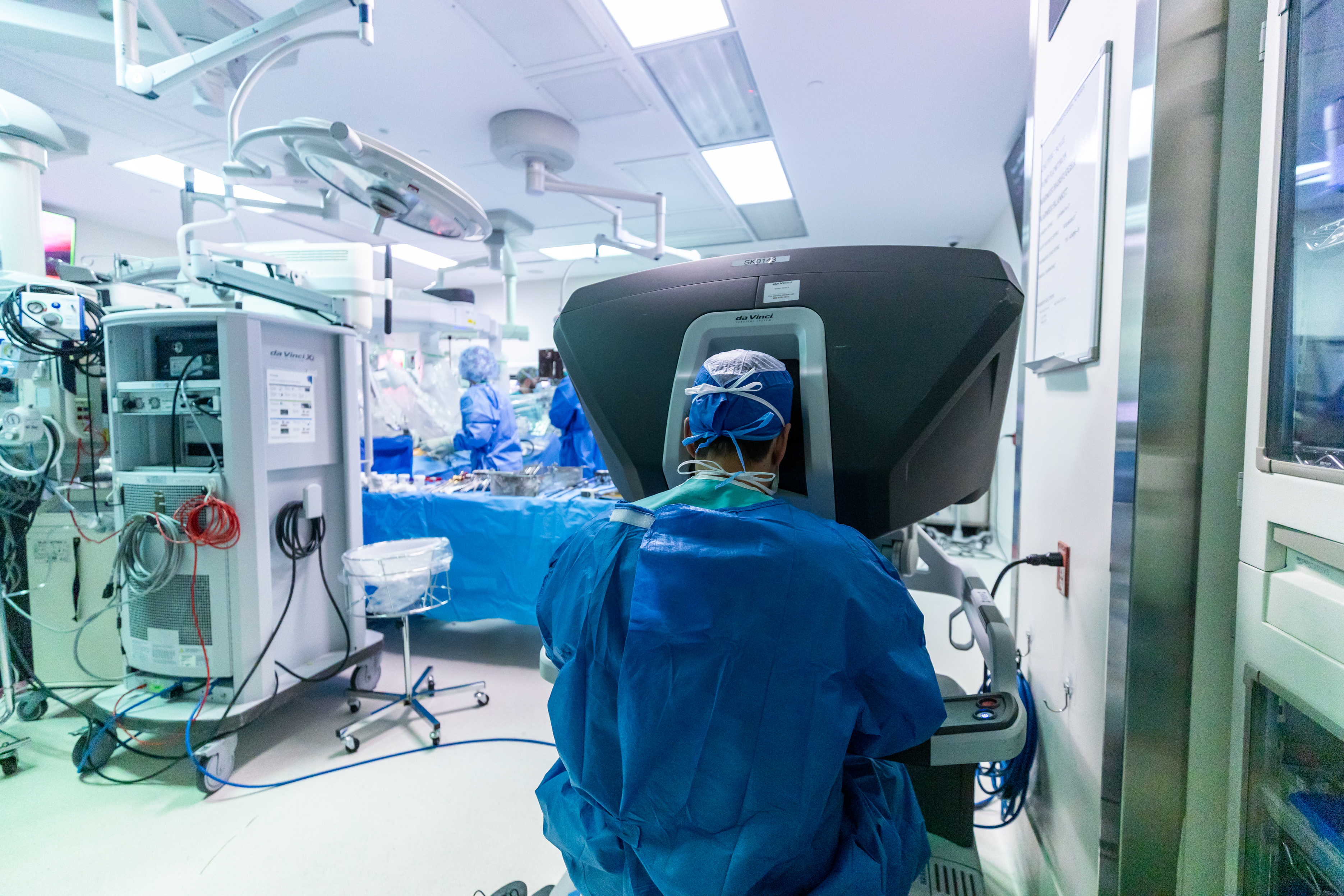 Operating room scene showing a surgeon in scrubs on the surgical robot in the foreground.