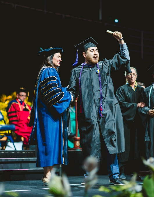 Graduate shaking President Nuñez's hand and celebrating with his diploma on the stage
