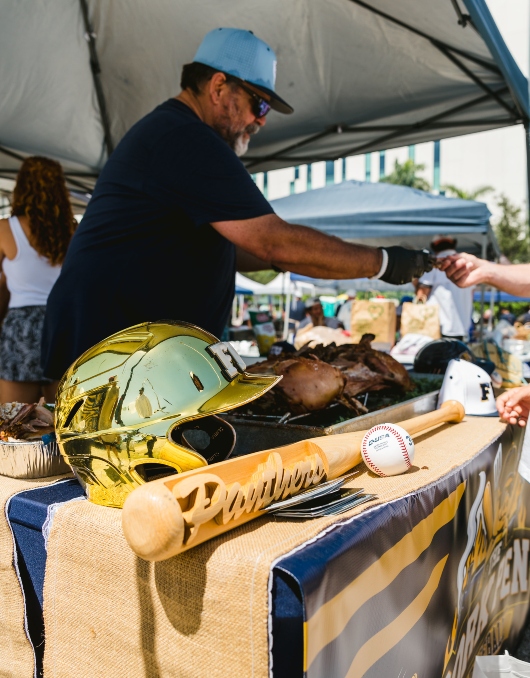 FIU Baseball pig roast team handing out pork, with baseball bat and helmet as props on the table